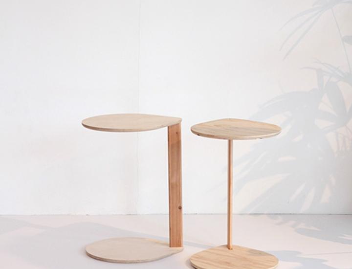 Deux tables d'appoint en bois clair du modèle Compass par Artifort. À gauche, une table avec une structure asymétrique en forme de C, et à droite, un modèle avec un pied central droit. Elles sont mises en situation dans un intérieur lumineux avec l'ombre d'une plante projetée sur le mur blanc en arrière-plan.