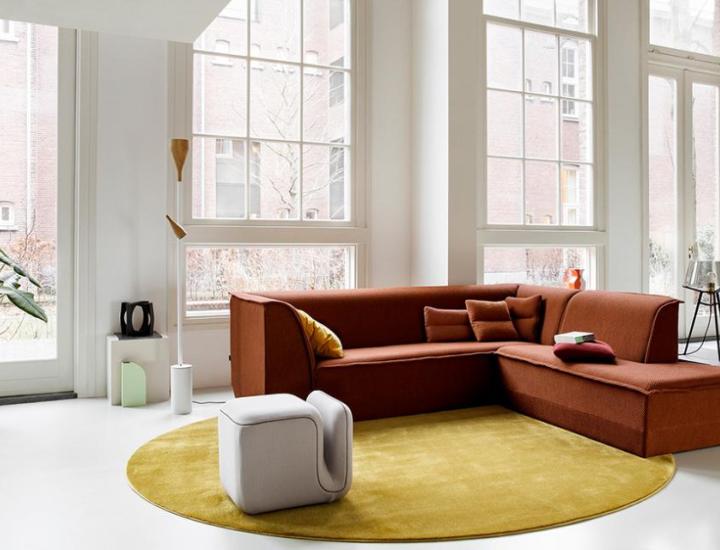 Mise en situation d'un pouf Moby d'Artifort en tissu gris clair, reconnaissable à sa fente formant une poignée. Il est posé sur un grand tapis rond jaune moutarde au centre d'une pièce très lumineuse. En arrière-plan, un vaste canapé d'angle couleur terracotta aménage cet espace moderne bordé de grandes baies vitrées blanches.