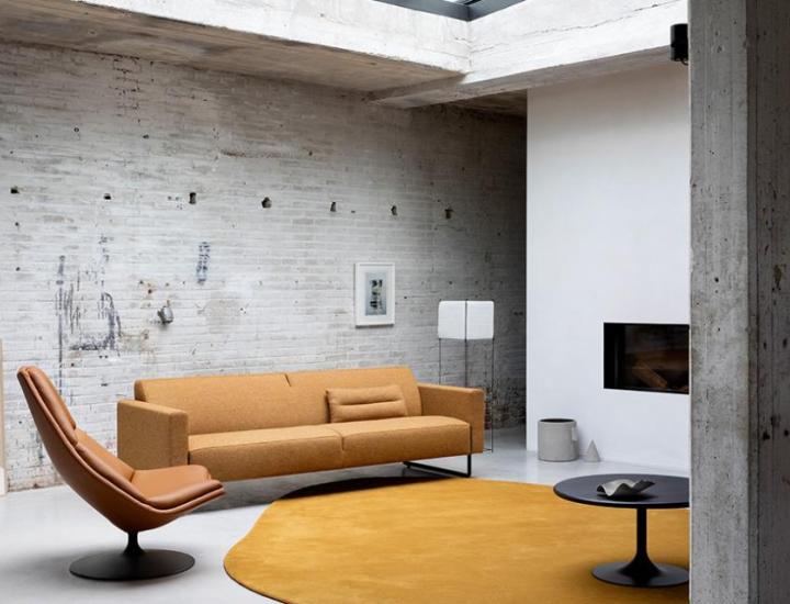 Aménagement de salon moderne de style industriel avec mur en briques blanches et sol en béton. Au premier plan, une table basse ronde noire Clarion Low d'Artifort posée sur un tapis jaune organique, près d'un canapé moutarde et d'un fauteuil pivotant en cuir camel. Une seconde table d'appoint Clarion noire est visible sur la gauche.