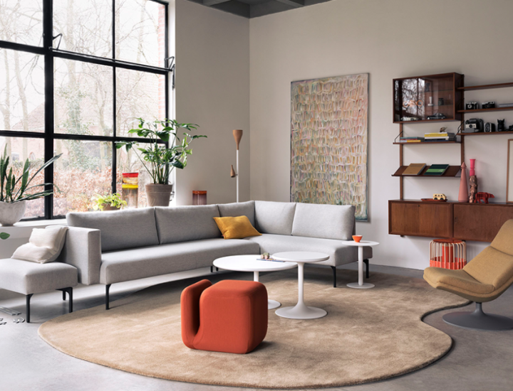 Grand salon lumineux au style contemporain avec une baie vitrée façon atelier. Au centre, sur un vaste tapis beige de forme organique, un ensemble de trois tables rondes blanches Clarion Low d'Artifort de différentes dimensions, entouré par un canapé d'angle gris clair, un pouf design orange et une étagère murale vintage en bois.