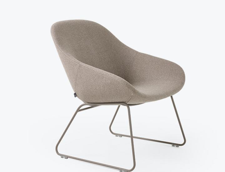 Un fauteuil moderne rembourré avec une coque d'assise et de dossier moulée et courbée, revêtu d'un tissu beige chiné. Il repose sur un piétement luge en métal de couleur bronze avec de petits patins. La vue est un angle de trois quarts bas, sur un fond de studio blanc.