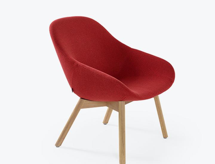 Un fauteuil moderne avec une coque d'assise et de dossier enveloppante, revêtu d'un tissu texturé de couleur rouge vif. Il repose sur un piétement classique composé de quatre pieds obliques en chêne clair, terminés par de petits patins. L'objet est présenté de trois quarts face sur un fond de studio blanc pur.